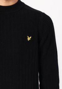 Maglione nero a coste con collo alto, caratterizzato da un piccolo logo di un'aquila gialla sull'area sinistra del petto. Motivo a maglia testurizzata su tutta la superficie.