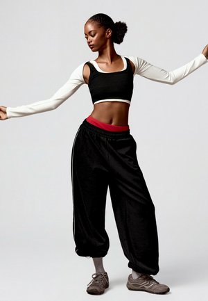 Jeune femme en pantalon noir ample, top court noir et blanc et baskets, pose avec les bras tendus devant un fond clair uni.