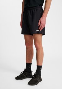 Schwarze Sportshorts mit elastischem Bund, Seitentaschen und einem kleinen Logo. Getragen mit schwarzen Sportschuhen und Socken mit einem Chevron-Muster.