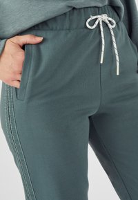 Teal joggers met een zachte textuur, voorzien van zijzakken, een koord in de taille en contrasterende witte stiksels aan de zijkanten.