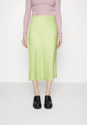 Kvinde iført en lysegrøn midilang nederdel, bleg lyserød langærmet croppet top og sorte platform slip-on sko, der står mod en hvid baggrund.