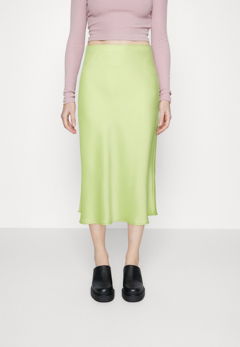 Femme portant une jupe midi vert clair, un top court à manches longues rose pâle et des chaussures plateformes noires à enfiler, debout devant un fond blanc.