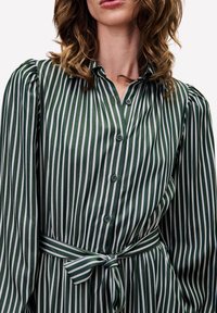 Robe verte à rayures fabriquée en tissu lisse, avec des rayures verticales blanches, un devant boutonné, un col et une ceinture à nouer à la taille.