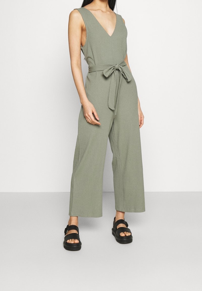 Femme portant une combinaison sans manches côtelée vert olive à jambes larges avec ceinture nouée, associée à des sandales noires à plateforme.