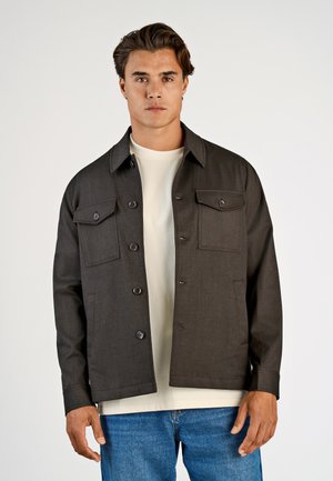 Donkergrijze button-up jas met een kraag, twee voorzakken en een relaxte pasvorm, over een lichtgekleurde T-shirt heen.