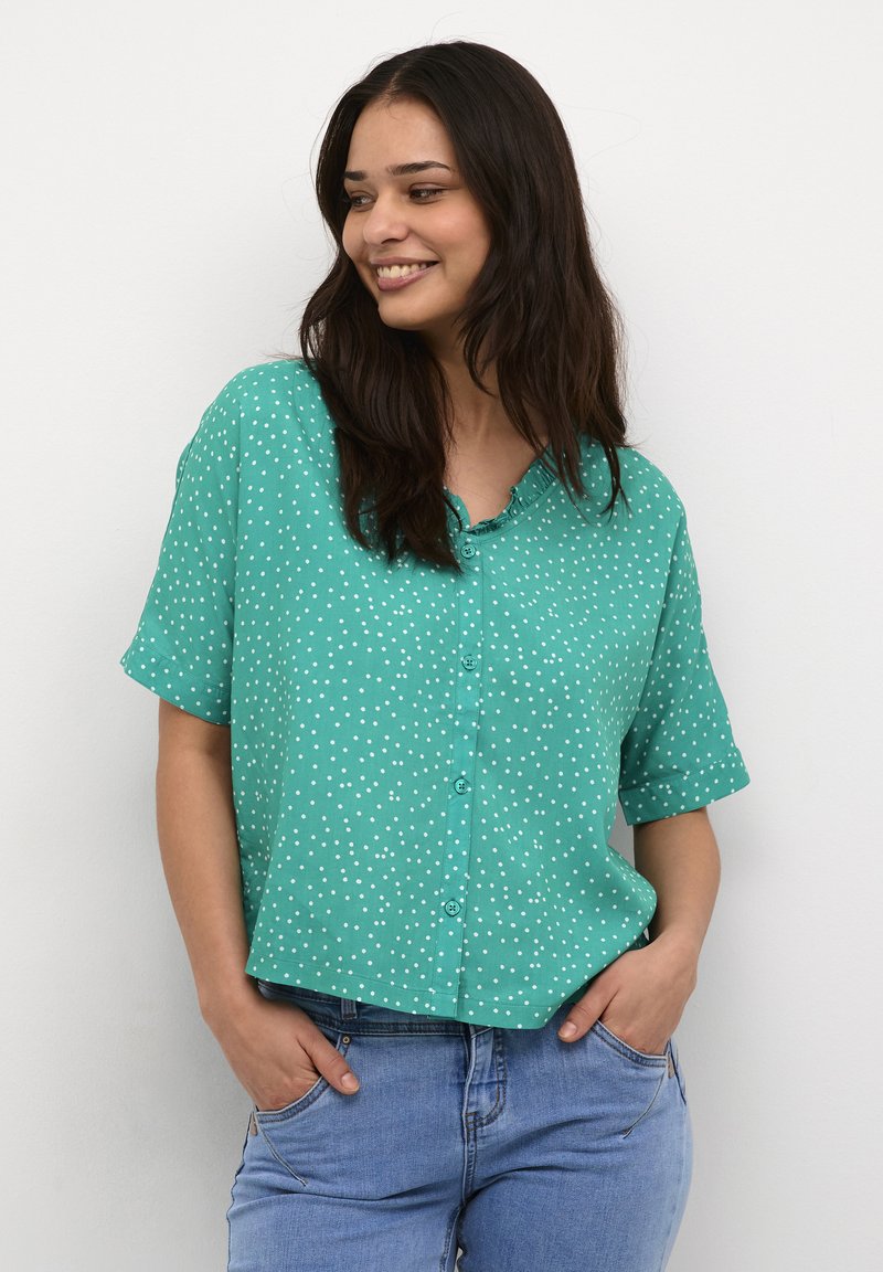 Camicetta turchese a maniche corte con pois bianchi, caratterizzata da una chiusura con bottoni e una vestibilità rilassata. Indossata con jeans blu e mani in tasca.