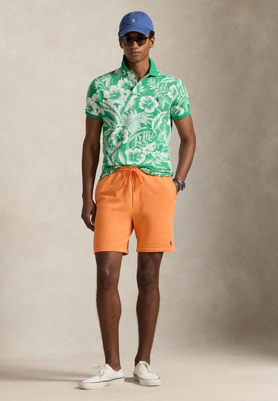 Πράσινο πόλο πουκάμισο με φλοράλ μοτίβο και γιακά, πορτοκαλί σορτς με κορδόνι, και λευκά αθλητικά παπούτσια, ολοκληρωμένα με μπλε καπέλο και γυαλιά ηλίου.