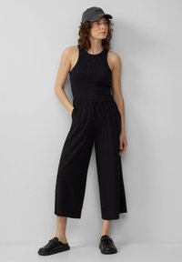 Zwart mouwloos topje met studs, gecombineerd met loszittende zwarte cropped broek en zwarte instappers, aangevuld met een grijze pet.