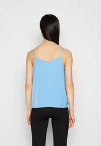 Camisola de cetim azul com alças finas, costas em forma de V e uma textura suave. Usada com leggings pretas.