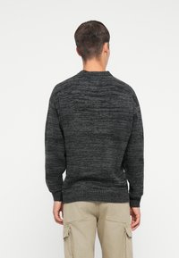Man som vänder ryggen mot kameran iförd en mörkgrå strukturerad tröja och beige cargobyxor mot en helt vit bakgrund.