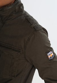 Olivgrön militärjacka i stil med hög krage, bröstficka och ett märke med Union Jack-design på ärmen.