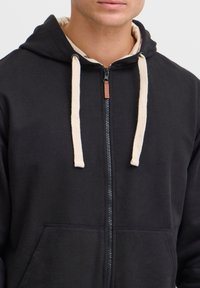 Sudadera con cremallera negra y forro color crema con cordones. Cuenta con bolsillos tipo canguro y una textura de tela suave.