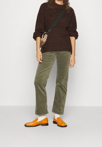 Personne portant un pull marron, un pantalon en velours côtelé vert, des chaussettes blanches, des mocassins orange et un sac bandoulière imprimé léopard sur un fond uni.