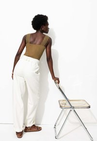 Débardeur vert olive avec des bretelles en dentelle ; pantalon large taille haute blanc ; tenant une chaise pliante transparente avec un cadre en chrome.