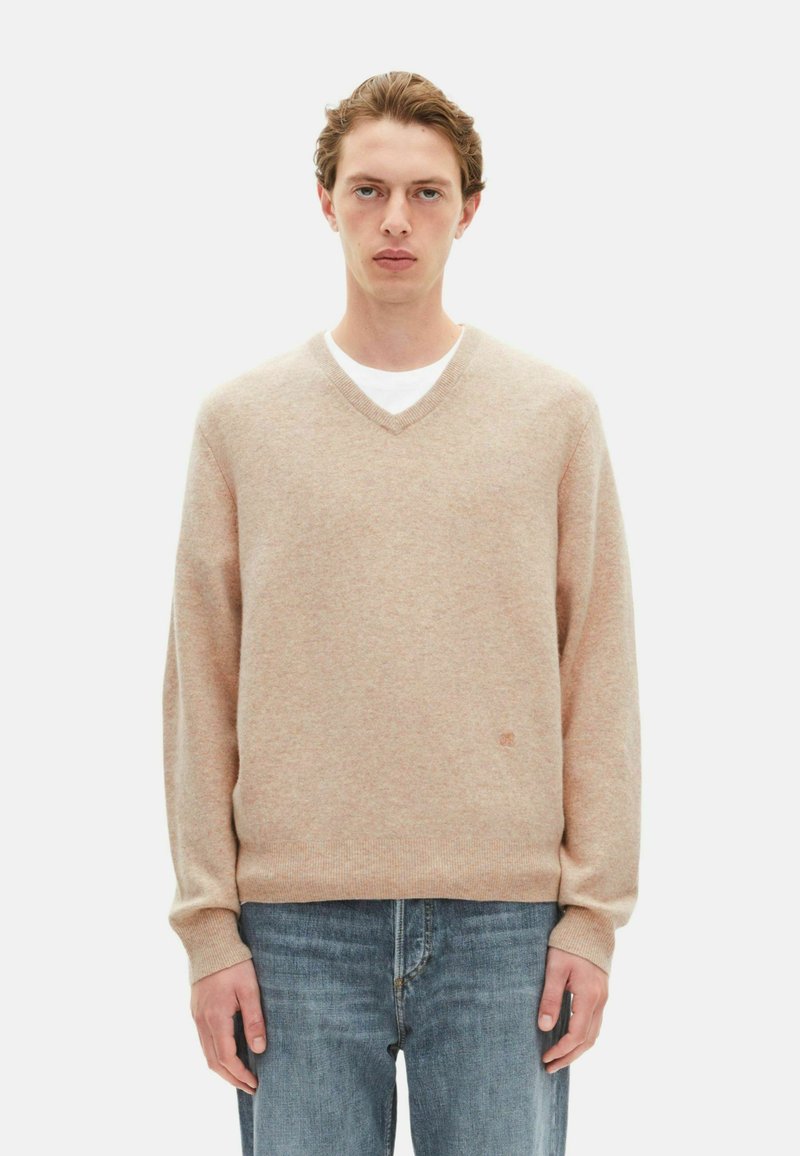 Suéter de punto beige con escote en V, fabricado en una tela suave, con mangas largas y un cuello acanalado, combinado con jeans de un azul claro. Detalle sutil del logo.