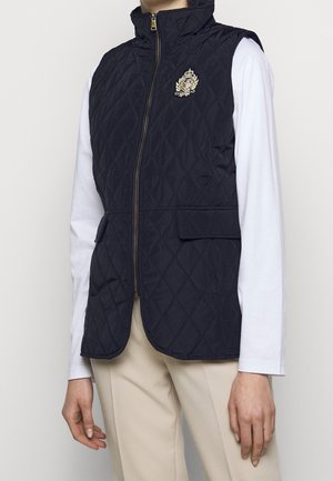 Personne portant un gilet matelassé sans manches bleu marine avec une fermeture éclair à l'avant, un blason brodé en or, une chemise blanche à manches longues et un pantalon beige.