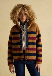 Svītrains flīsa žakets brūnā, tumši zilā un bordo krāsā ar augstu apkakli. Aprīkots ar snap pogām un mīkstu tekstūru. Nēsāts virs baltas krekla.