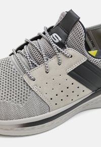 Chaussure de sport grise avec dessus en mesh, accents en daim, détails perforés et semelle en caoutchouc texturée. Équipements noirs contrastants.