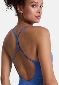 Μπλε μαγιό με λεπτούς λουριά στο πίσω μέρος σε σχήμα σταυρού, κατασκευασμένο από απαλό ύφασμα, με χαμηλή πλάτη και μινιμαλιστικές λεπτομέρειες.