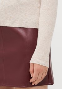 Beige neule, jossa on teksturoidu pinta ja ribatut hihansuut, yhdistettynä burgundinpunaisiin nahkahousuihin, jotka ovat sileäpintaiset ja siistit.