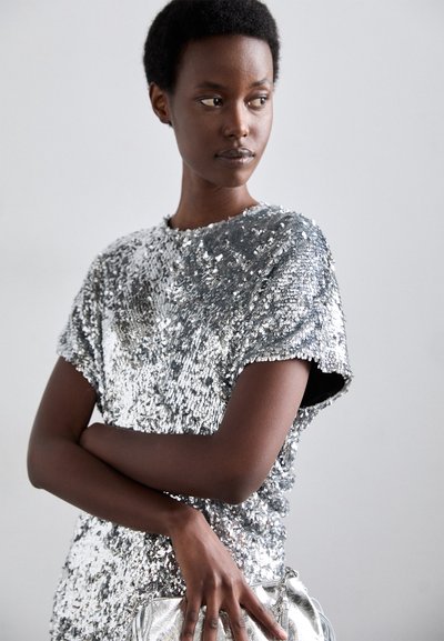 Silver sequin top with short sleeves, round neckline, and loose fit, paired with a shiny silver handbag. Smooth surface with reflective textures.