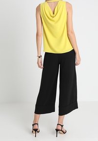 Femme portant un haut jaune sans manches avec un dos drapé, un pantalon noir large et court, des sandales noires à talons hauts, et une montre-bracelet.