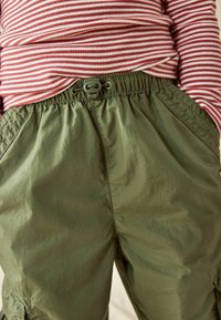 Grüne Cargoshorts mit elastischem Bund und Kordelzugdetails, kombiniert mit einem rot-weiß gestreiften Langarmshirt.
