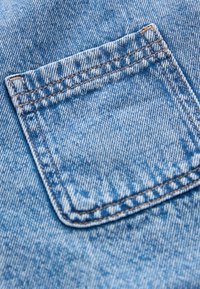 Šviesiai mėlynos džinsinės medžiagos su lopinės kišene. Matomi siuvimo siūlės kontrastingos rudos spalvos. Faktūra atrodo dėvėta su subtiliais raštais.