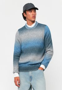 Pull en tricot dégradé de nuances de bleu et de gris, col rond, manches longues, texture douce, associé à une chemise blanche à col et une casquette.