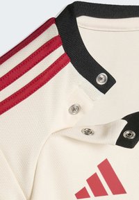 Weißes, strukturiertes Sporttrikot mit schwarzem Kragen, roten drei Streifen an der Schulter, Druckknöpfen und einem teilweisen roten Markenlogo sichtbar.