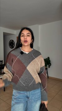 Tânără cu păr lung și închis la culoare, purtând un pulover cu model argyle și blugi, vorbind într-o cameră în care se văd un ceas și o plantă.