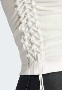 Witte blouse met lange mouwen, gekenmerkt door een gerimpelde textuur langs de zijnaad en een trekkoord aan de onderrand. Glad materiaal met een lichte glans.