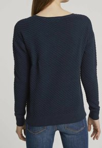 Pull en maille texturée bleu marine au fit décontracté, col rond et poignets côtelés. Motif gaufré visible sur le tissu. Associé à un denim.