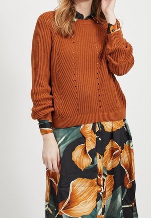 Frau trägt einen rost-orangefarbenen Strickpullover über einem schwarzen Kleid mit großem orange-grünem Blumenmuster, steht vor einem schlichten Hintergrund.