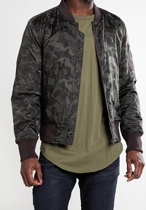 Uomo che indossa una giacca bomber mimetica verde scuro sopra una t-shirt lunga verde oliva e pantaloni neri, in piedi con le braccia rilassate.