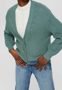Sweter kardigan w stonowanej zieleni z głębokim dekoltem w kształcie V, z zapięciem na guziki i prążkowanymi mankietami; noszony na białej, fakturowanej bluzce i zestawiony z niebieskimi dżinsami.