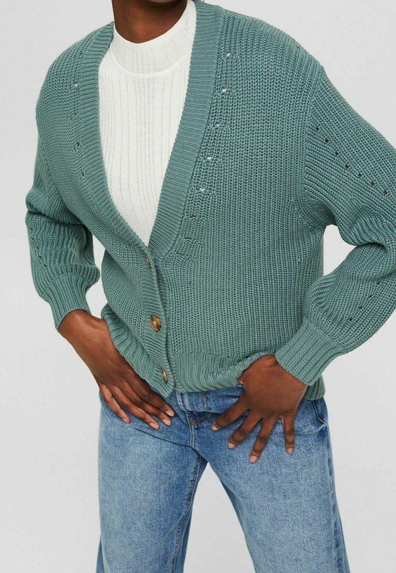 Sweter kardigan w stonowanej zieleni z głębokim dekoltem w kształcie V, z zapięciem na guziki i prążkowanymi mankietami; noszony na białej, fakturowanej bluzce i zestawiony z niebieskimi dżinsami.