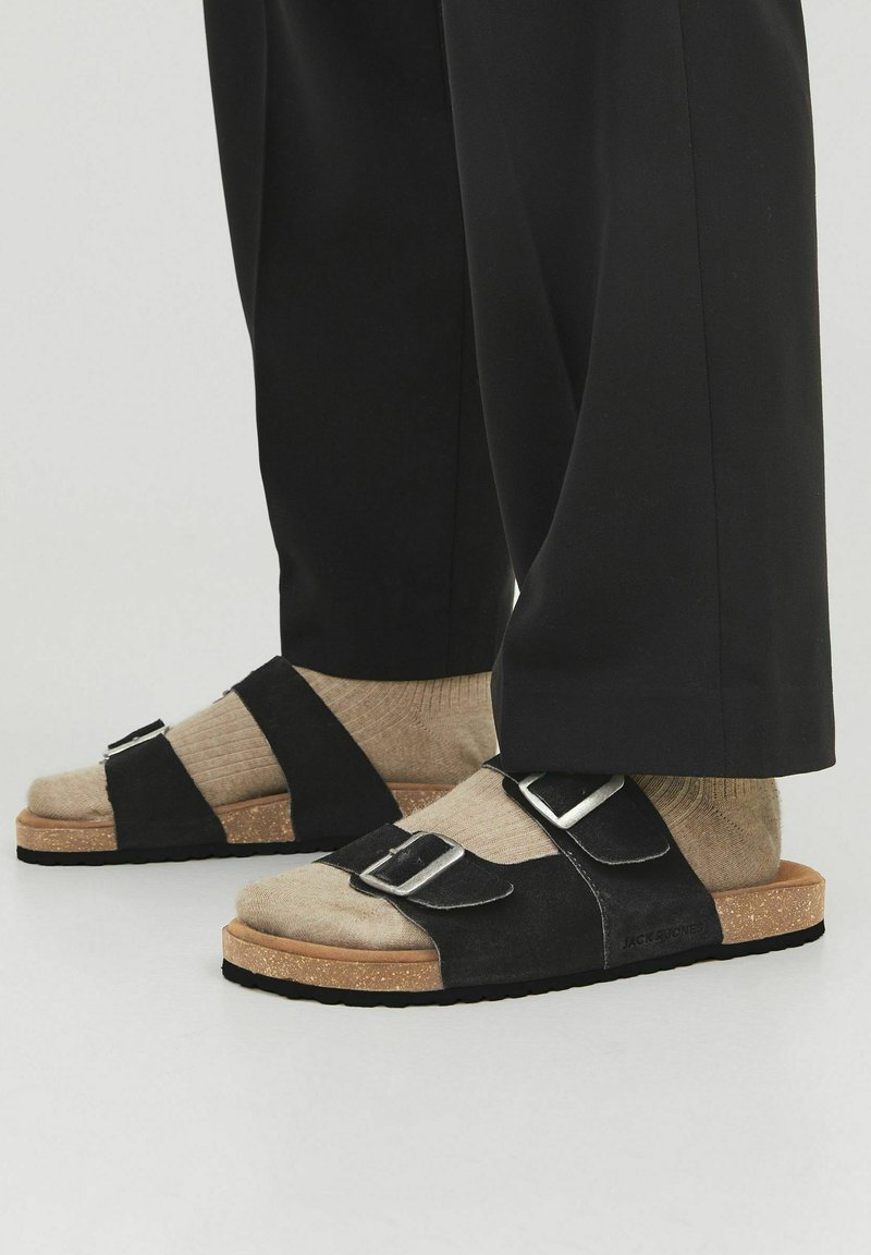 Sandales noires àDouble bride avec boucles en argent, semelle en liège texturé, portées avec des chaussettes beiges claires. Caractéristiques d'un design plat et d'un orteil arrondi.