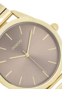 Montre-bracelet dorée avec un boîtier rond et un bracelet, arborant un cadran dégradé marron, des index horaires fins et un design minimaliste.