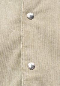 Tissu en velours côtelé beige avec des rainures verticales, doté d'un bouton-pression argenté. Texture douce visible, mettant en valeur le design du matériau.