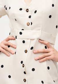 Mains d’une femme sur les hanches portant une robe beige à pois noirs, boutons marron et ceinture en tissu assortie à la taille.