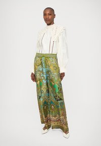 Witte blouse met kanten kraag strak om de nek; groene en blauwe patroonwijdte broek met een elastische taille en trekkoord; witte schoenen.