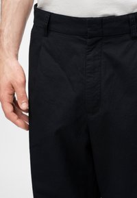 Homme portant un pantalon noir taillé sur mesure et une chemise blanche, main détendue sur le côté, taille et partie supérieure de la jambe visibles.