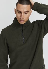 Pullover in pile verde scuro con colletto alto e zip a metà, caratterizzato da una texture morbida e maniche lunghe, con dettagli di branding minimali.