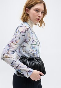 Γυναίκα με κοντά κόκκινα μαλλιά που φοράει ένα διαφανές λουλουδάτο τοπ και μπεζ ζιβάγκο, κρατώντας μια μαύρη δερμάτινη τσάντα φάκελο με πτυχώσεις.