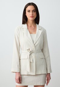 Blazer in lino beige chiaro con righe verticali, design a un bottone, due tasche frontali e cintura in tessuto. Abbinato a una gonna coordinata.