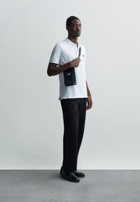 Férfi fehér pólót, fekete nadrágot és fekete cipőt viselve áll, és egy fekete keresztpántos táskát tart egy sima szürke háttér előtt.