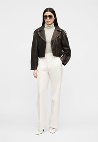 Žena u smeđoj kožnoj jakni, prugastom golfu, bijelim hlačama visokog struka, bijelim salonkama i sunčanim naočalama, stoji na jednoj običnoj pozadini.