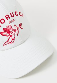 Bela kapica iz blaga s rdečim vezenim besedilom in grafiko cheruba z lokom in srcem. Struktura z obliko z ukrivljenim robom.