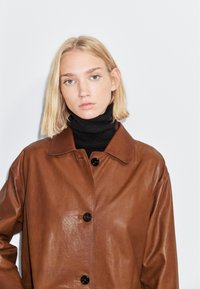 Bruine leren jas met een knopenfront, spitse kraag en een gladde textuur. Draagt over een zwarte coltrui. Eenvoudig ontwerp, geen patronen.